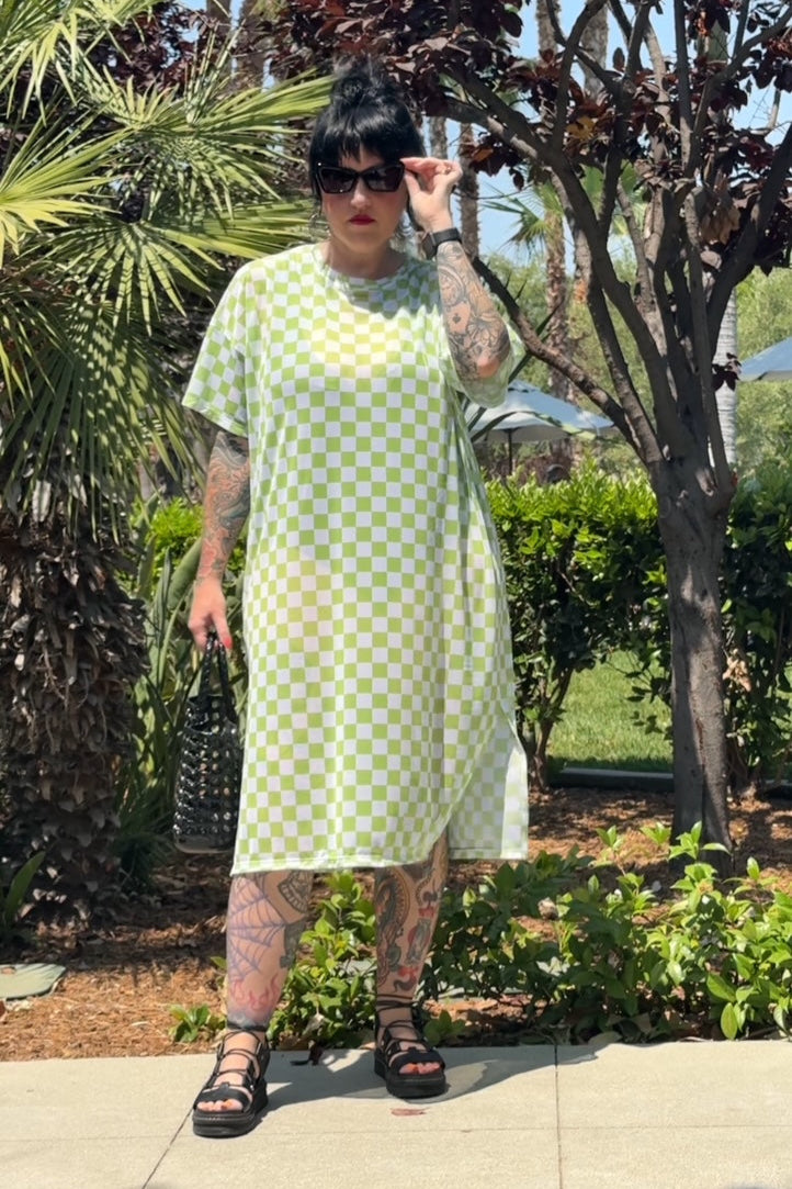 Person wearing a green checkered dress standing outdoors with trees and plants in the background
