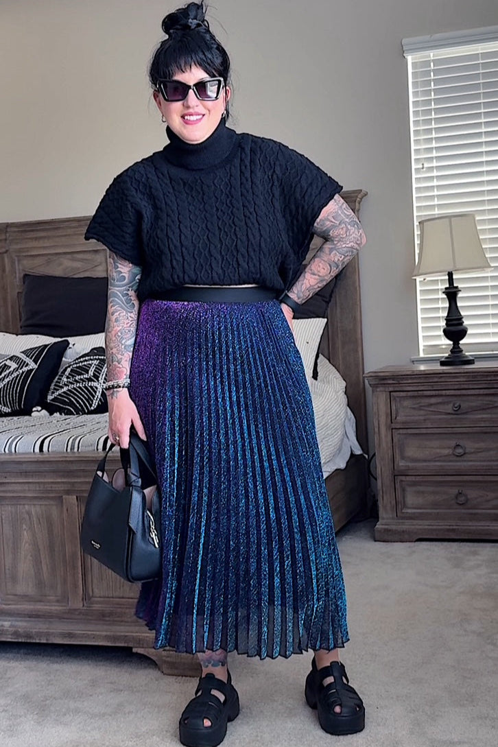 Person wearing a black turtleneck and blue pleated skirt in a bedroom setting.