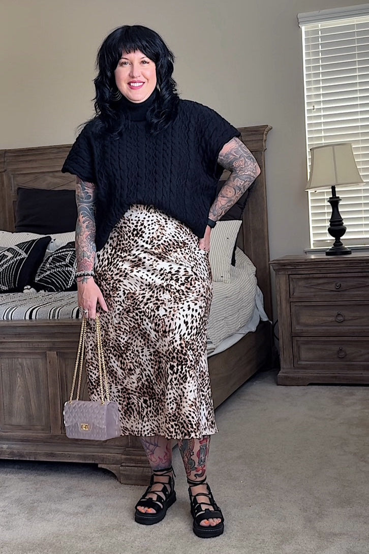 Woman standing in a bedroom wearing a black top and leopard print skirt.