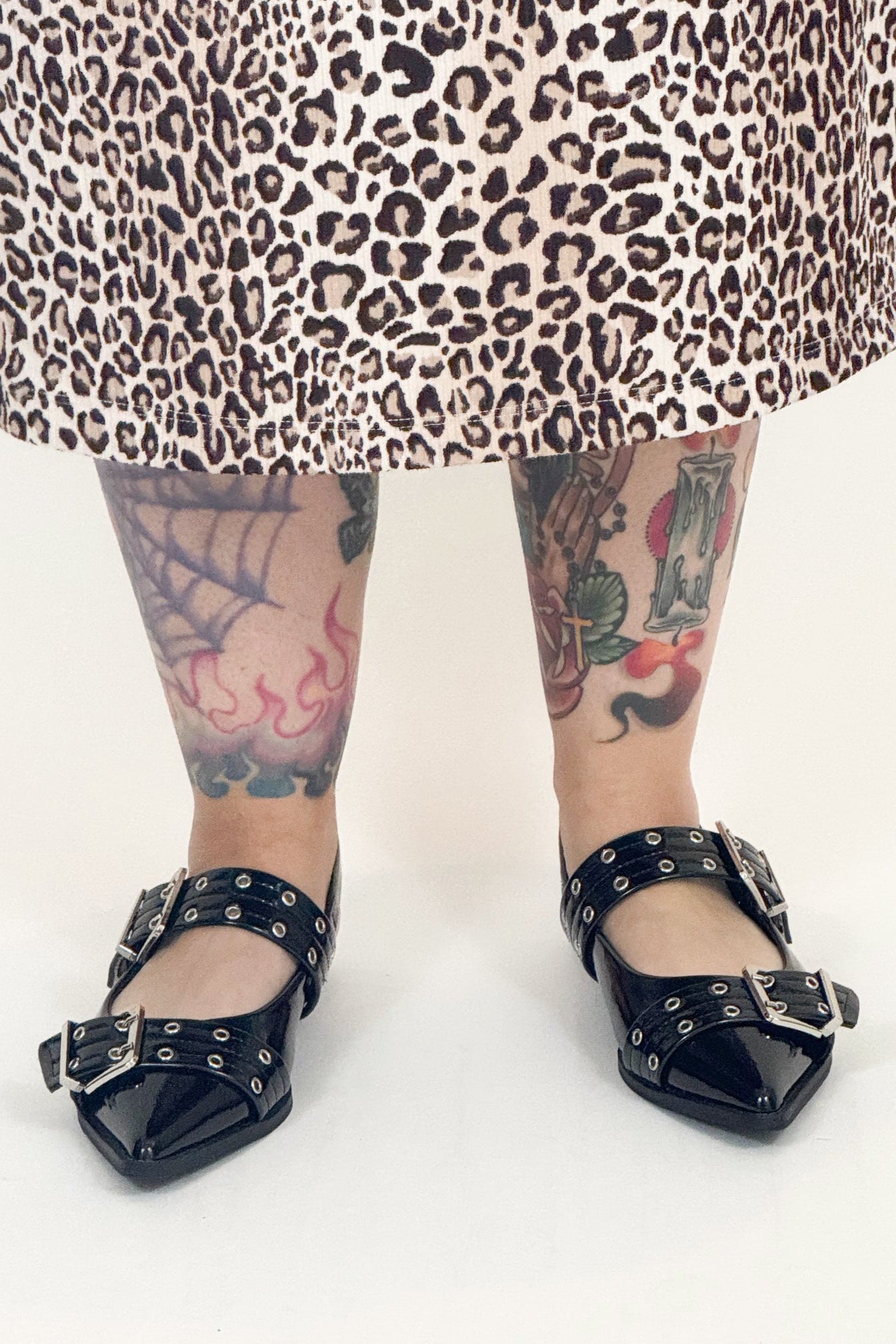 Person wearing black studded shoes with a leopard print skirt and tattoos on a white background