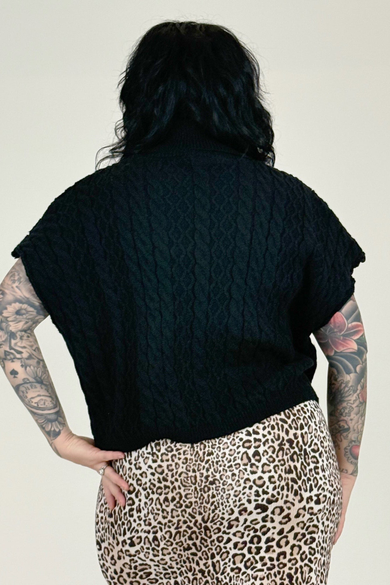 Woman wearing a black cable knit vest over a leopard print skirt on a plain background