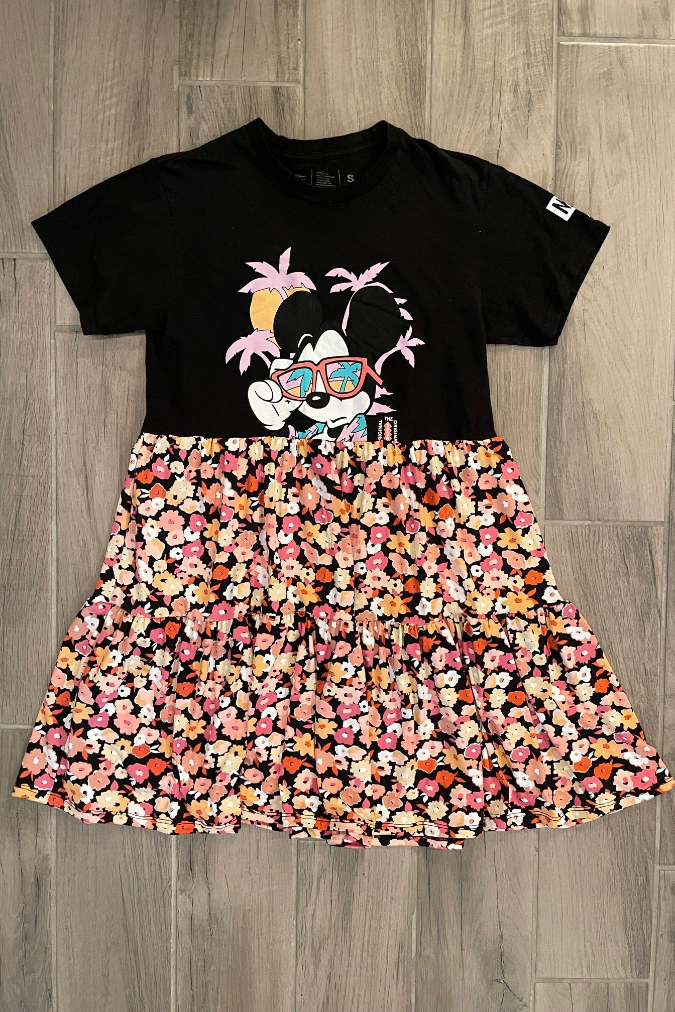 A t-shirt dress with a black floral design on the top and a multicolored floral print on the skirt, displayed on a wooden floor.