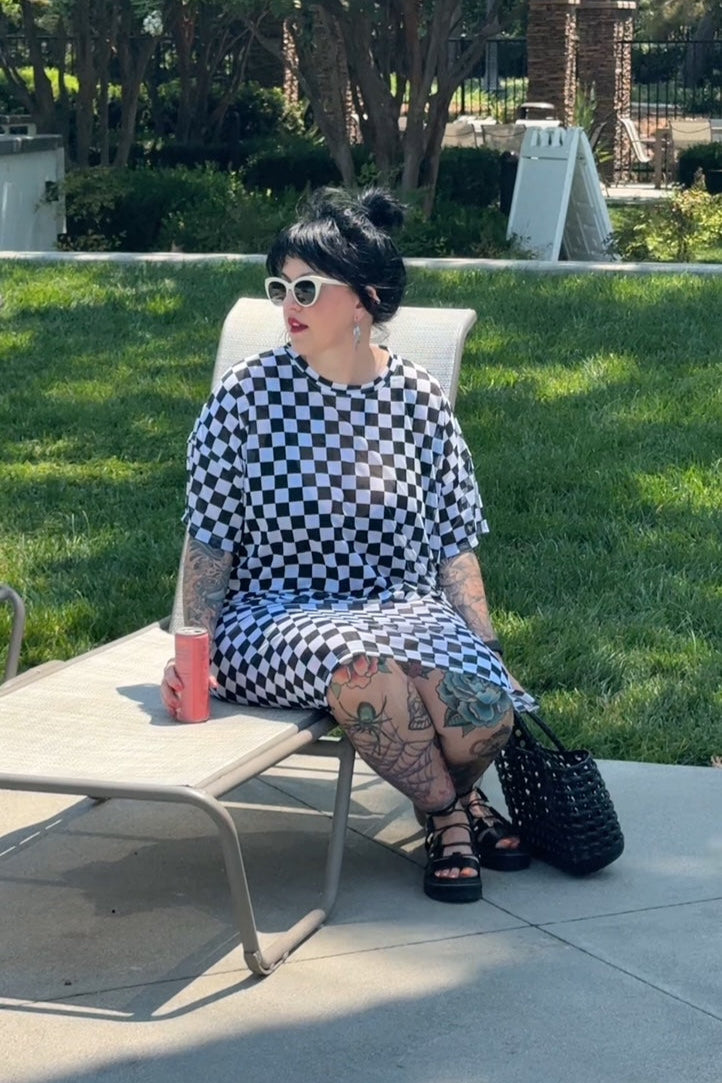 Woman sitting on a chair outdoors, wearing a checkered dress and sunglasses.