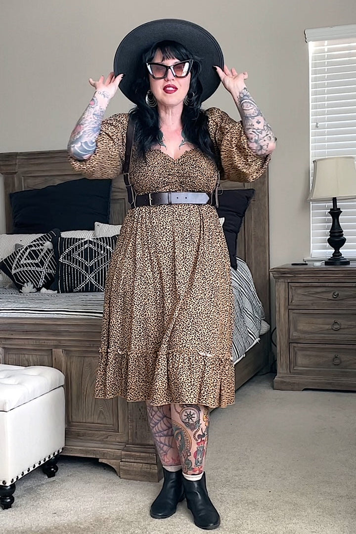 A person wearing a brown leather fashion harness with an adjustable waist belt and shoulder straps, featuring metal buckles and ring. The harness is styled with a leopard print dress, black hat, and sunglasses.