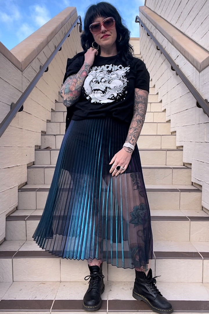A person wearing a pleated skirt with iridescent turquoise and purple hues, paired with a black top and standing on stairs.