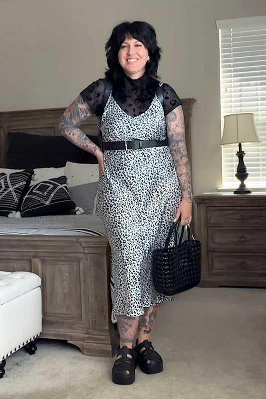 A person wearing a sleeveless, spaghetti strap, midi-length dress with a leopard print design, paired with a black belt, shoes, and carrying a black bag.