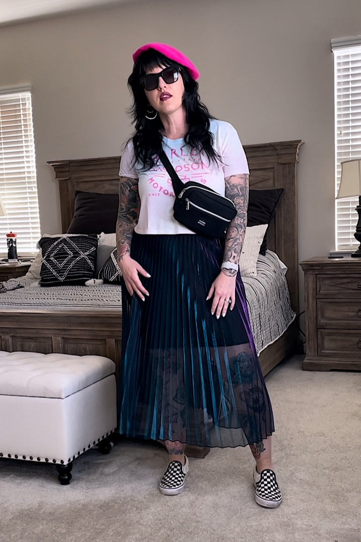 A person wearing a midi-length pleated skirt with iridescent turquoise and purple colors, paired with a white top and pink hat, standing in a room with a bed and sunglasses on a surface in the background.