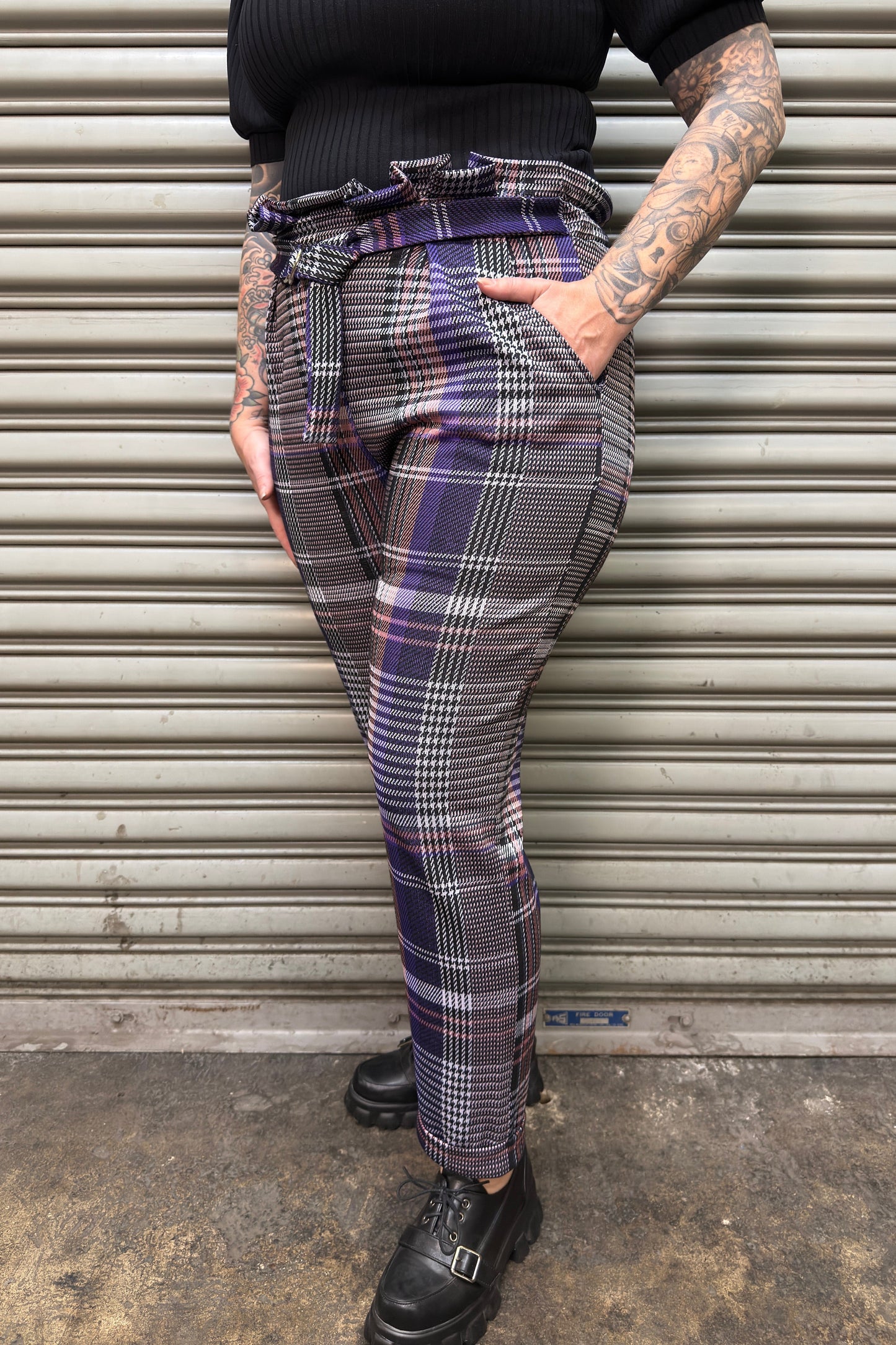 Person wearing plaid pants standing against a metal shutter.