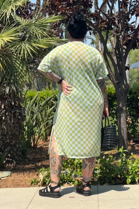 Person wearing a green checkered dress standing outdoors with plants in the background