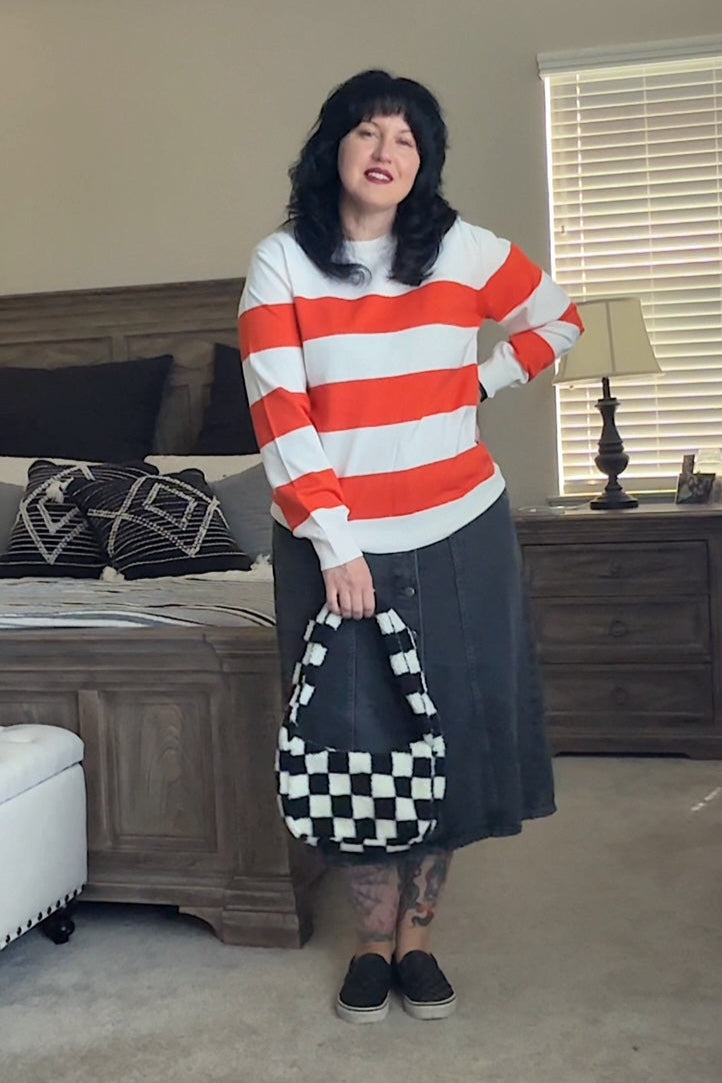 Person wearing a red and white striped sweater and black skirt holding a checkered bag in a bedroom.