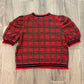 A red and black plaid short-sleeve shirt laid flat on a gray wood-look tile floor.