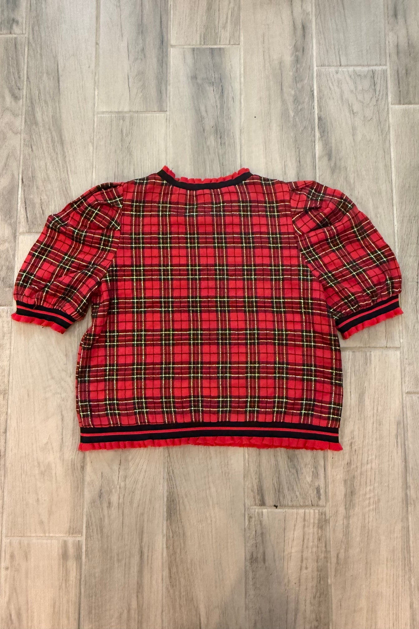 A red and black plaid short-sleeve shirt laid flat on a gray wood-look tile floor.