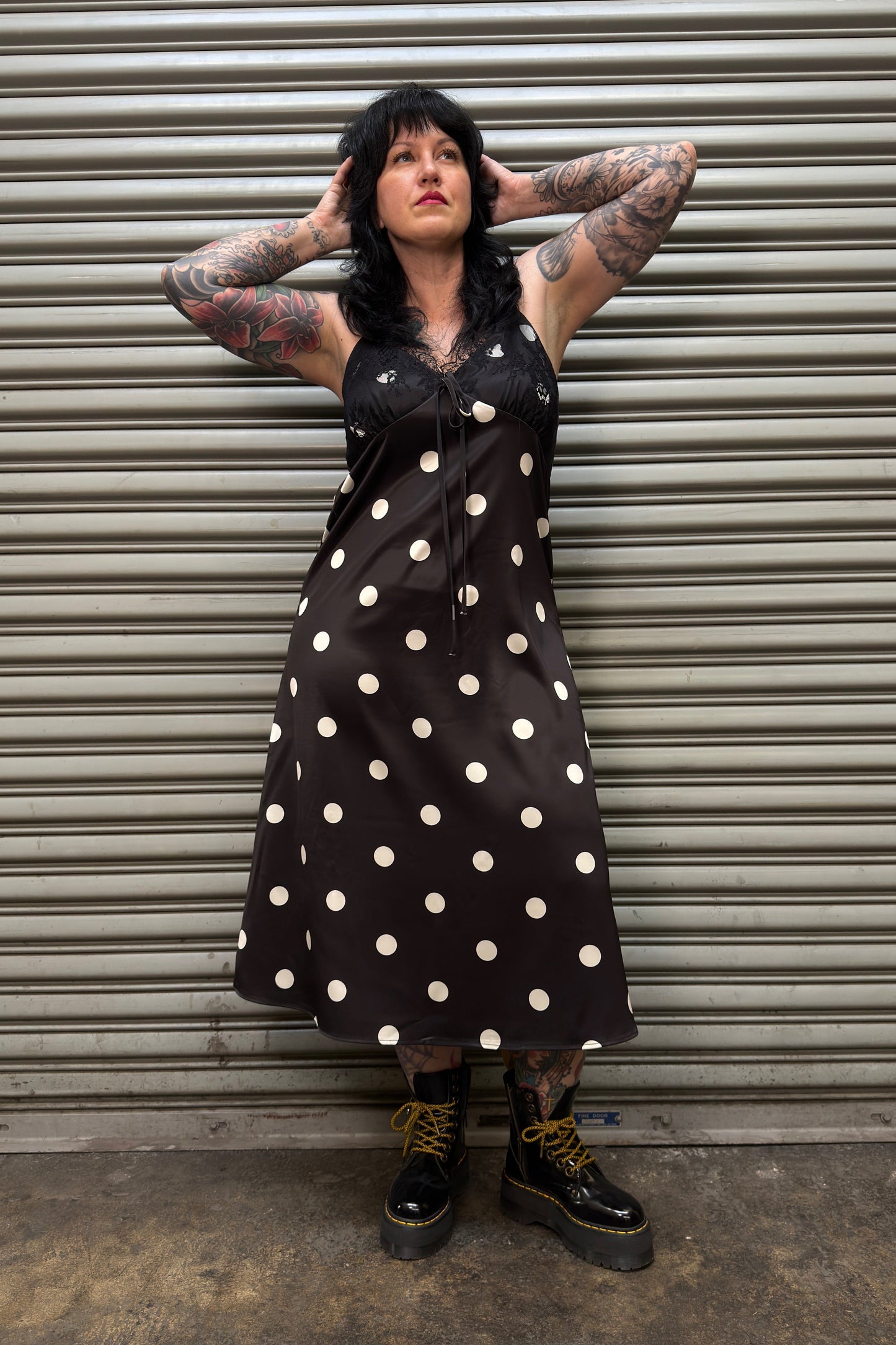 Person wearing a black polka dot dress standing against a metal shutter.