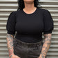 Person wearing a black ribbed top and high-waisted jeans with tattoos on arms against a metallic shutter background