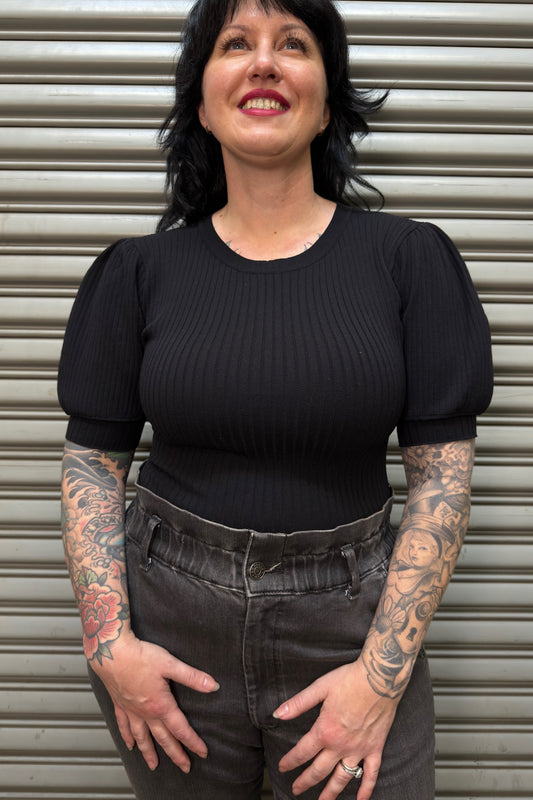 Person wearing a black ribbed top and high-waisted jeans with tattoos on arms against a metallic shutter background