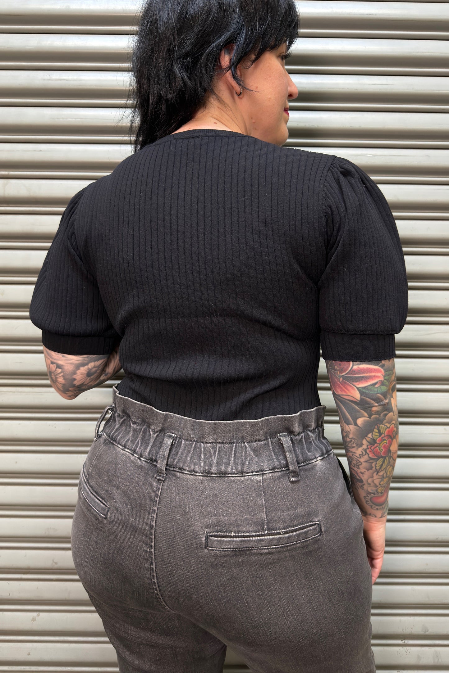 Person wearing a black ribbed top and gray pants standing in front of a metallic shutter.