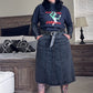 Person wearing a black sweatshirt with graphic design and a denim skirt in a bedroom setting.