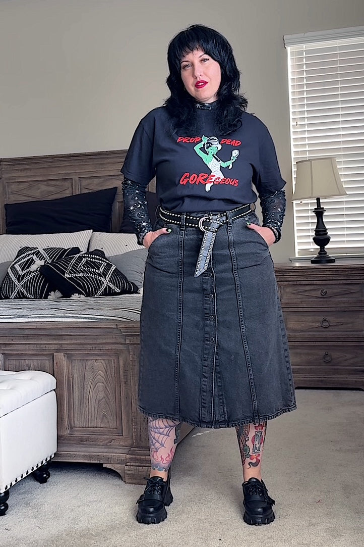 Person wearing a black sweatshirt with graphic design and a denim skirt in a bedroom setting.