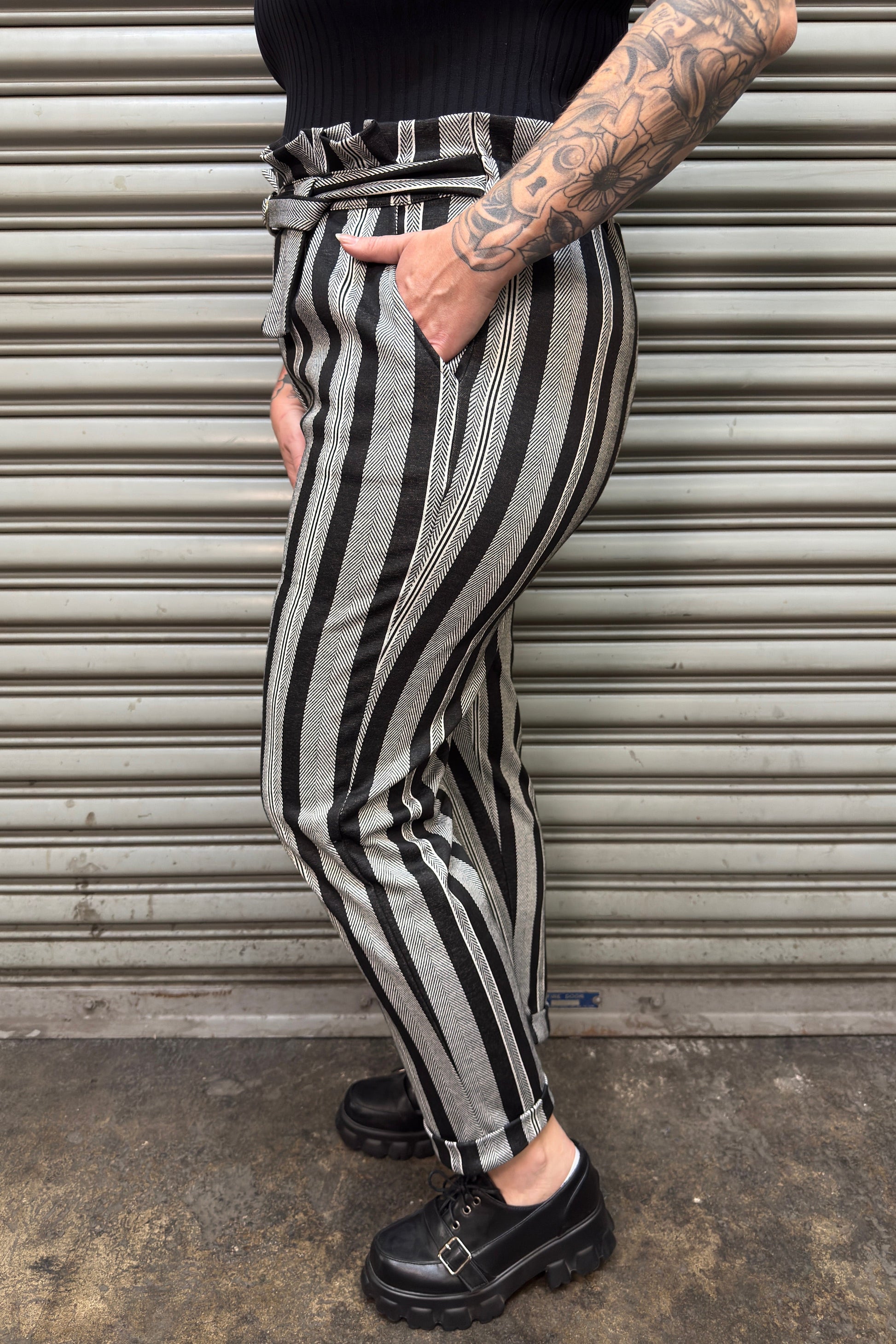 Person wearing black and white striped pants with tattoos visible, standing against a metallic shutter.