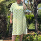 Person wearing a green checkered dress standing outdoors with trees and plants in the background