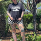 Person wearing a black t-shirt with a graphic design and checkered shorts standing outdoors.