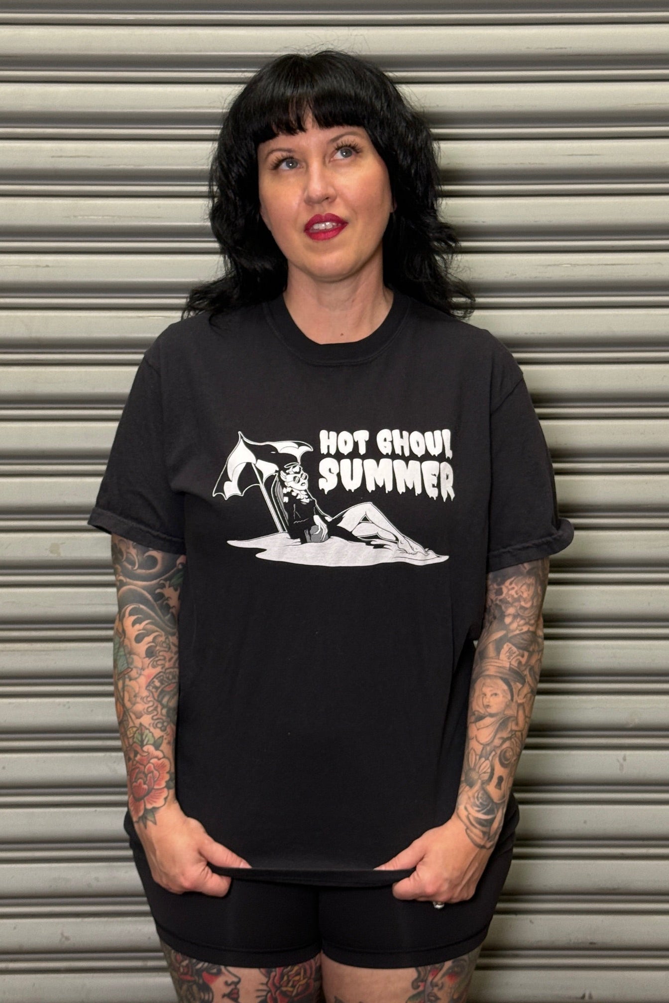 Person wearing a black t-shirt with graphic design and text, standing against a metallic shutter.
