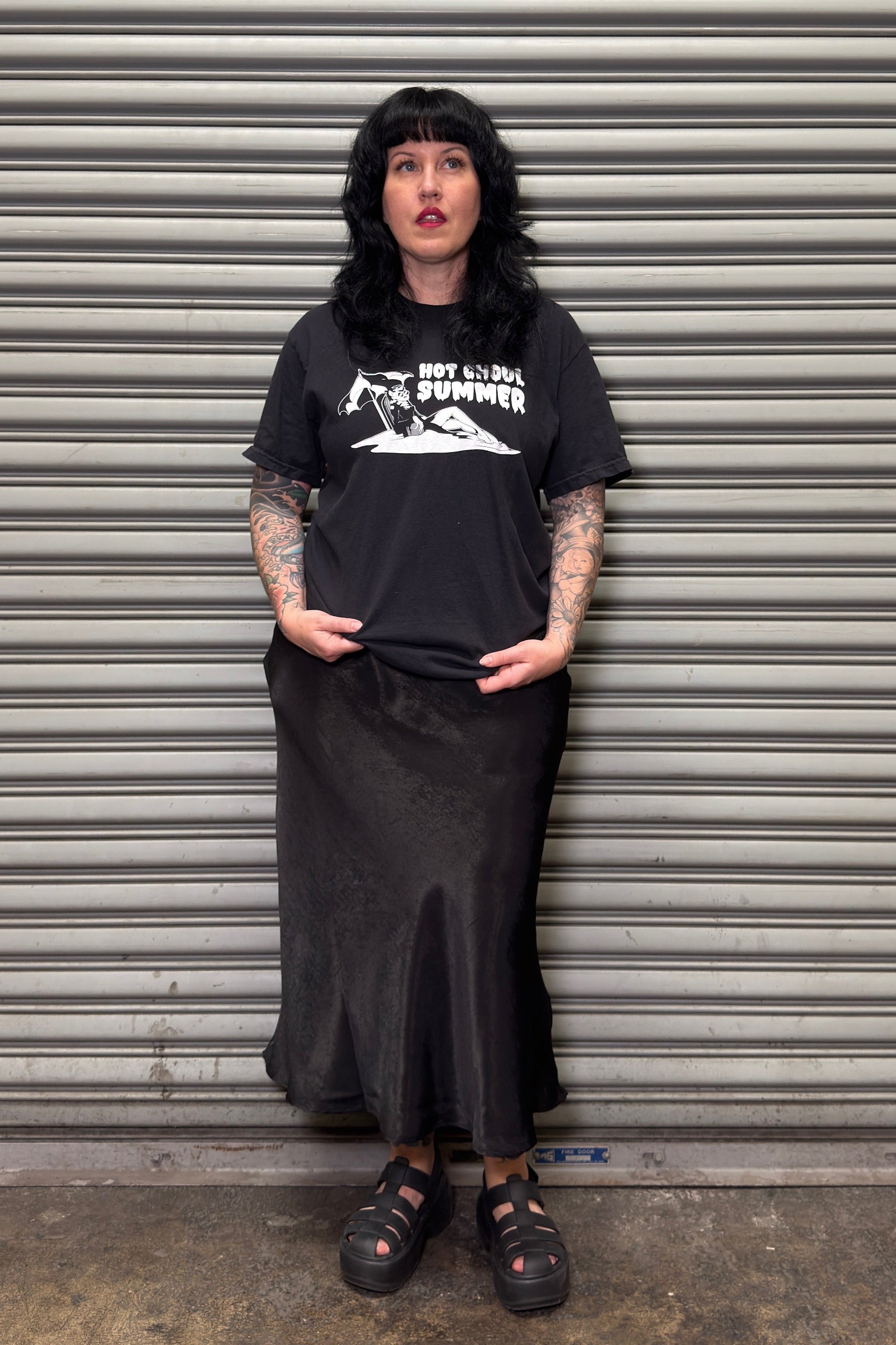 Person wearing a black t-shirt and skirt standing in front of a metal shutter.