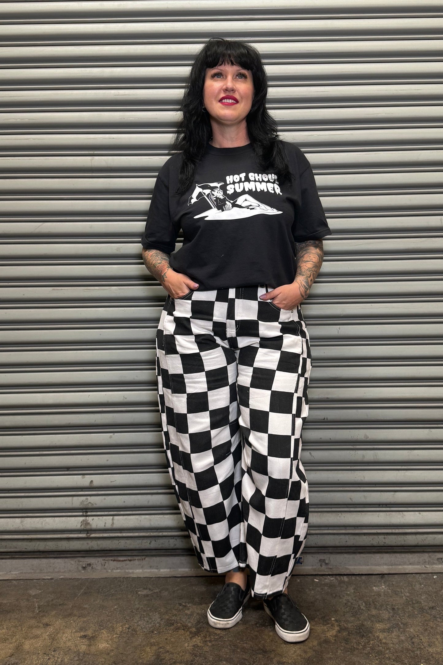 Person wearing a black t-shirt with graphic design and checkered pants standing in front of a metallic shutter.