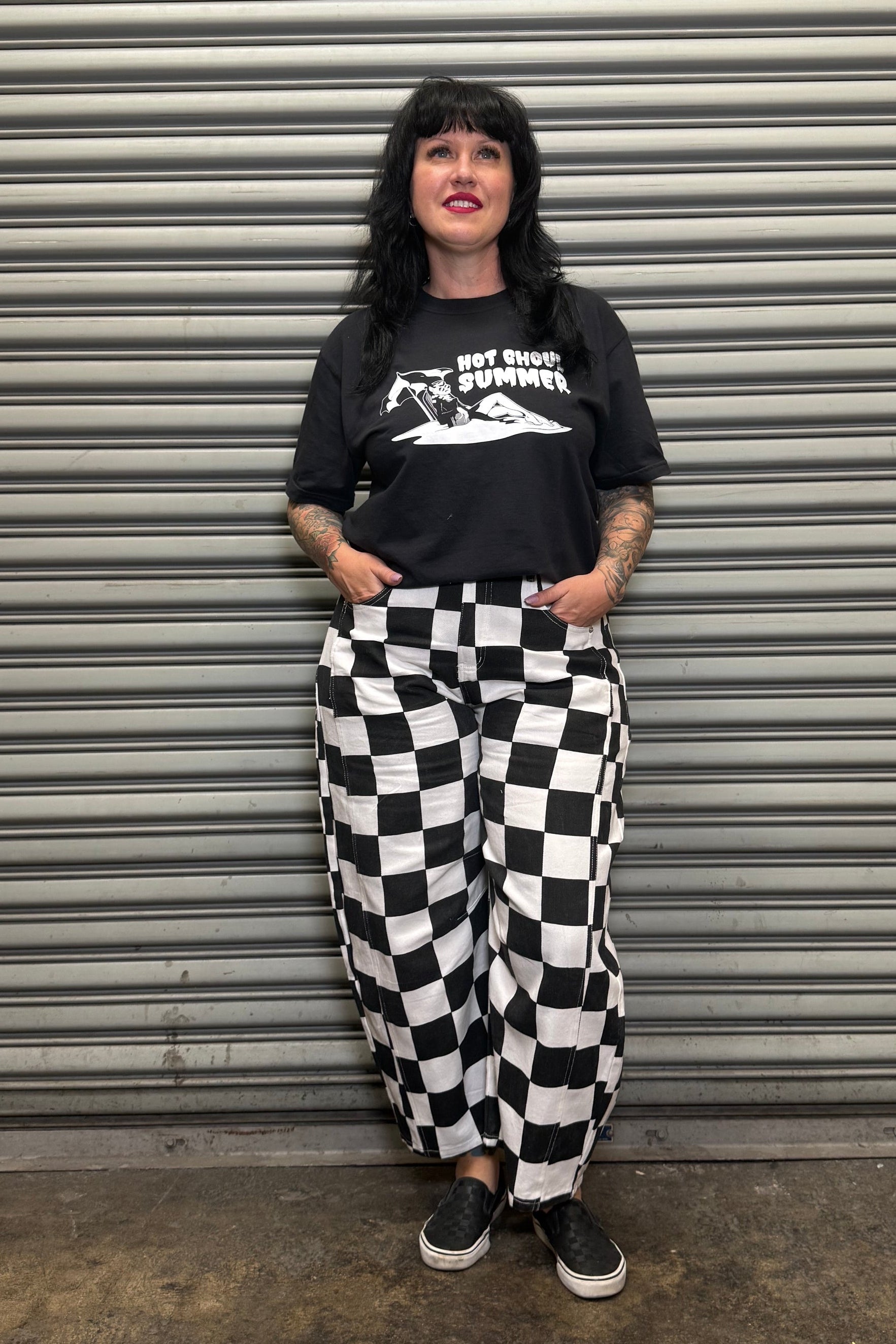 Person wearing a black t-shirt with graphic design and checkered pants standing in front of a metallic shutter.