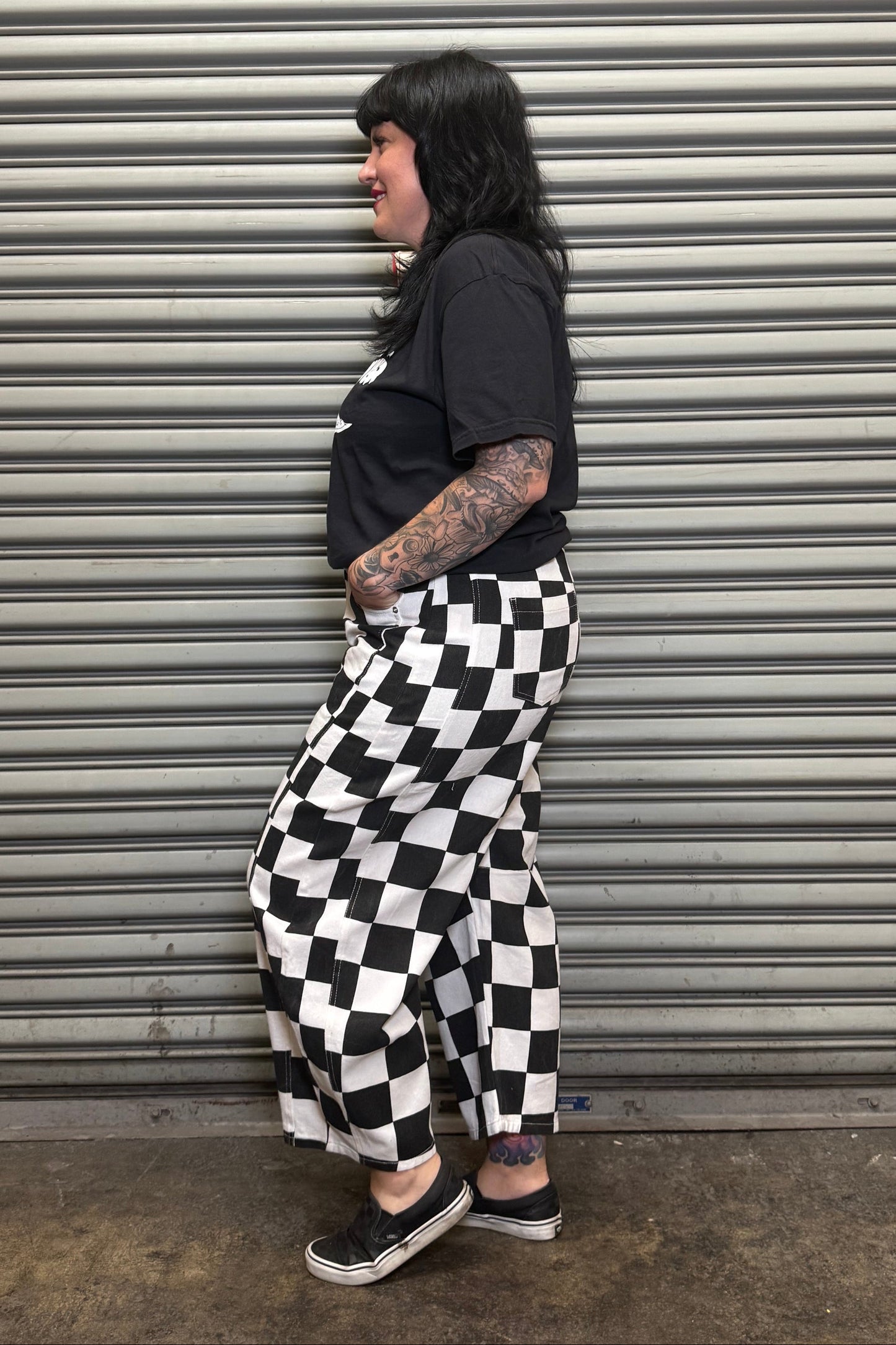Person wearing a black t-shirt and checkered pants standing against a metal shutter.