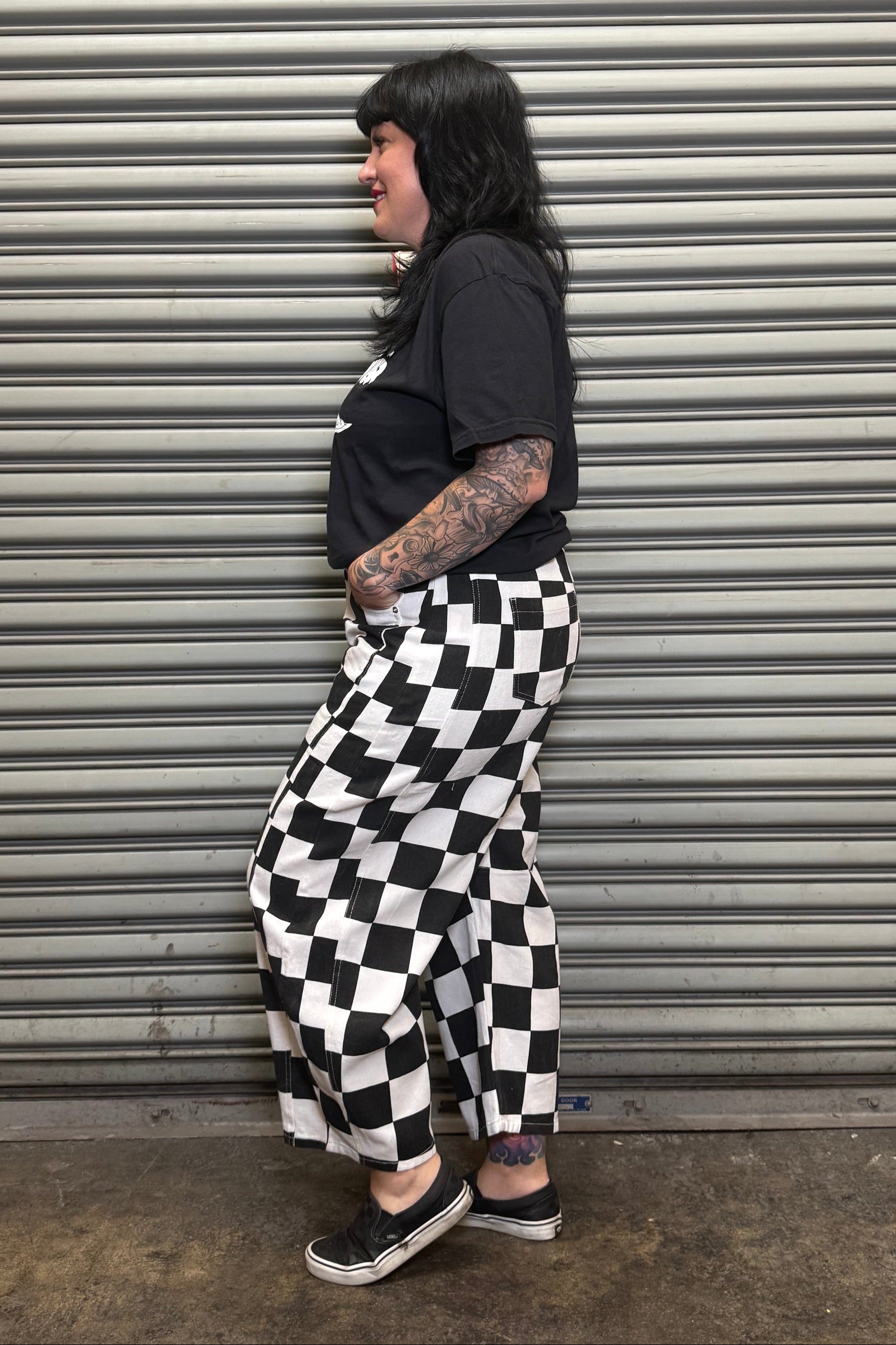 Person wearing a black t-shirt and checkered pants standing against a metal shutter.