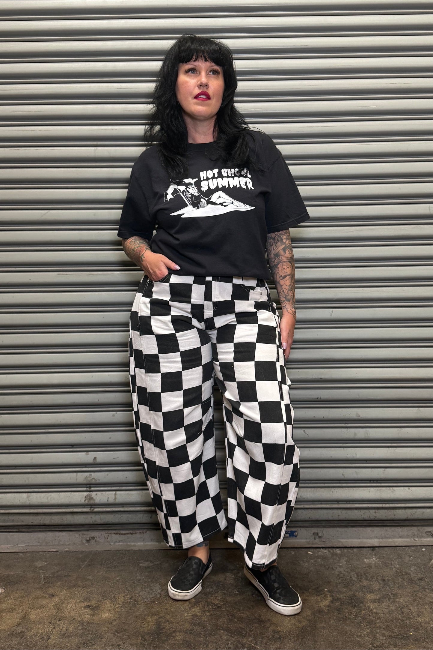 Person wearing a black t-shirt with white graphics and checkered pants standing in front of a metal shutter.