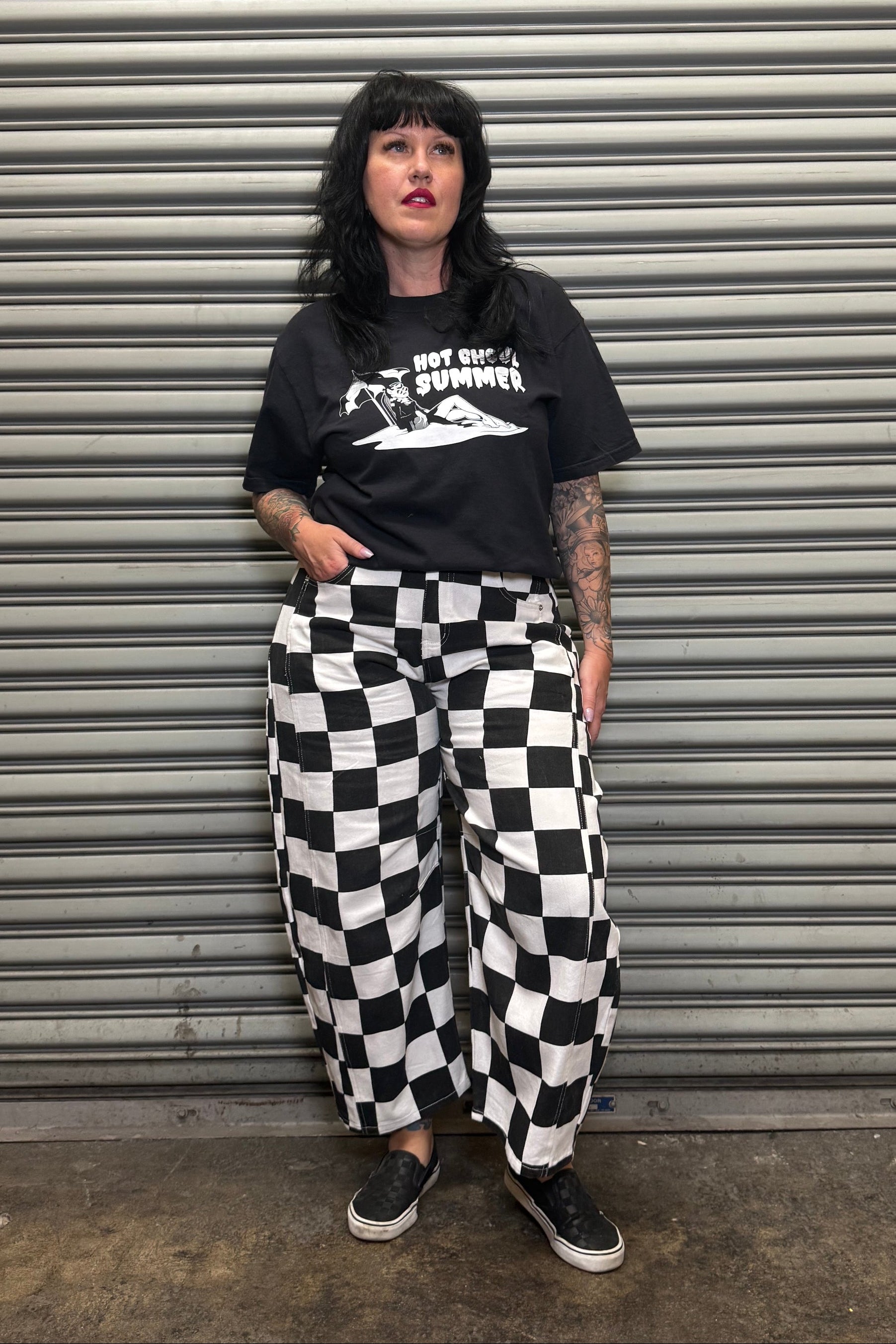 Person wearing a black t-shirt with white graphics and checkered pants standing in front of a metal shutter.