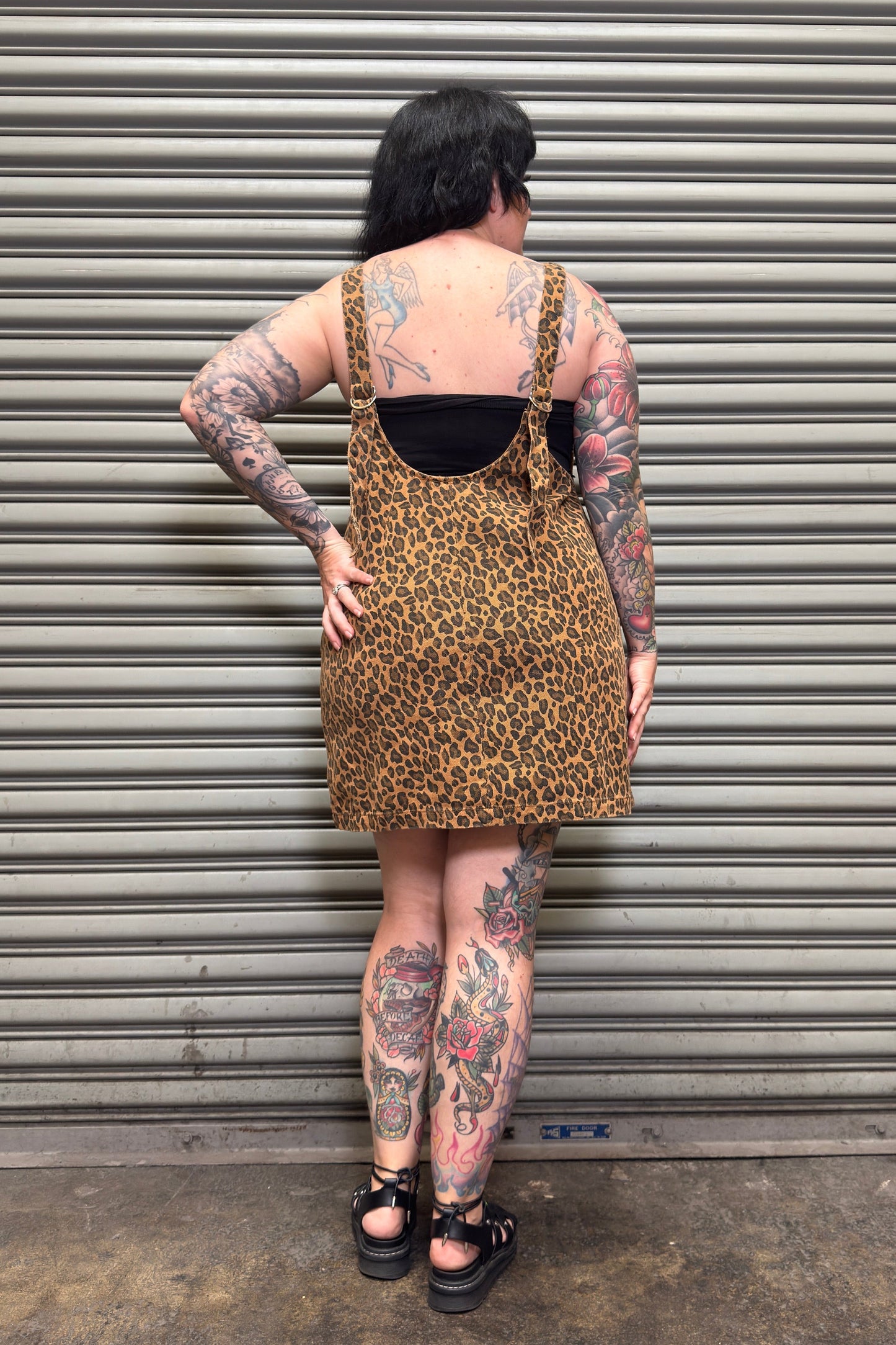 Person wearing a leopard print dress with tattoos standing against a metal shutter.