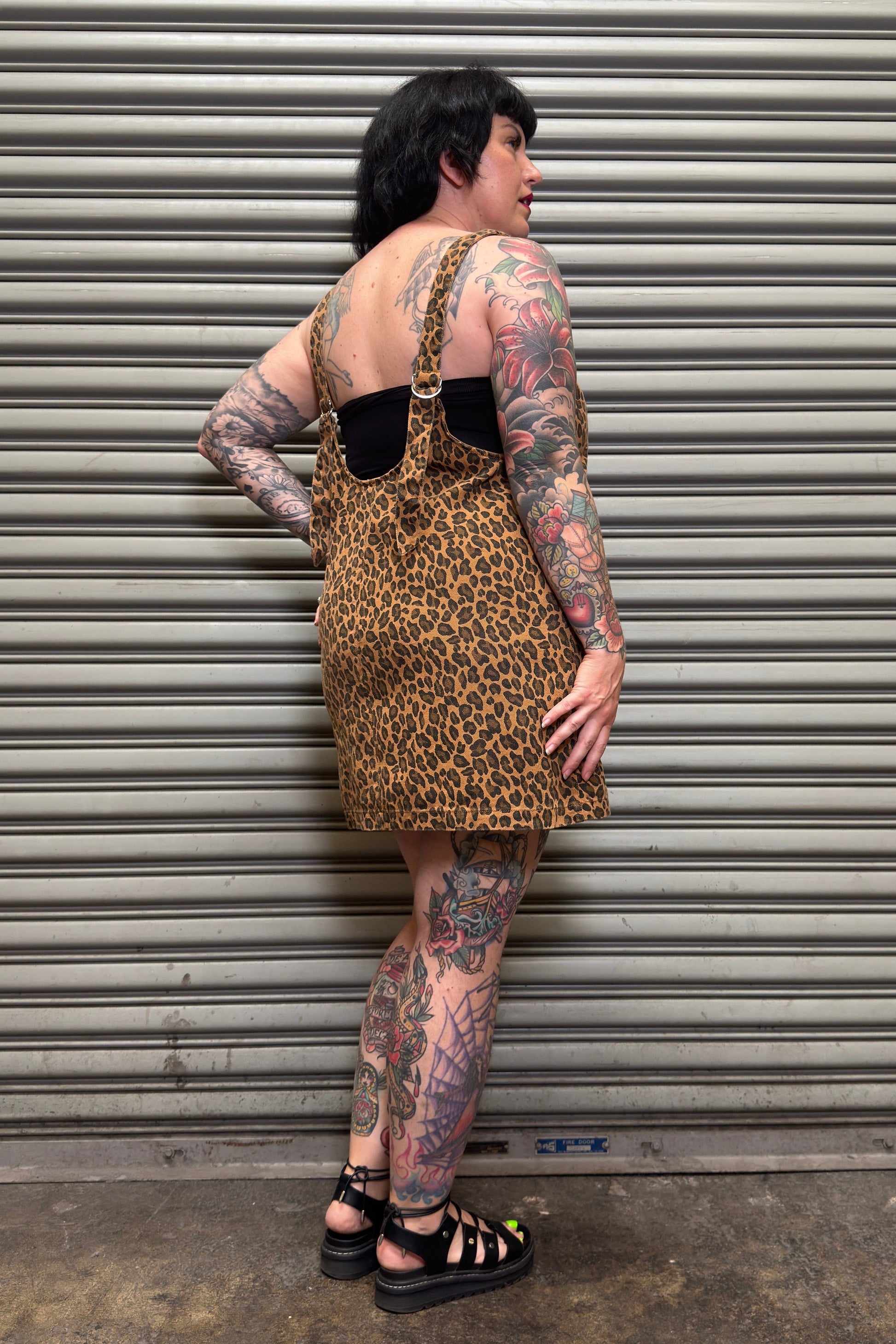 Person wearing a leopard print dress with black overalls against a metal shutter background