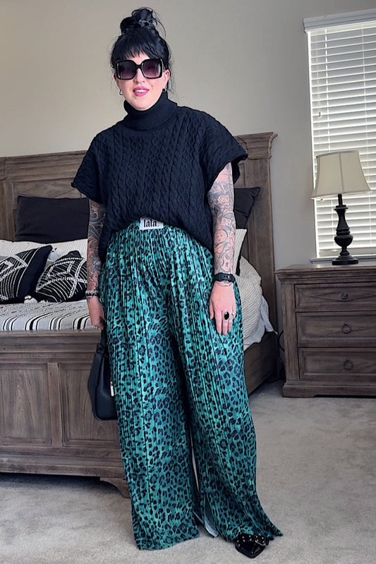 Woman wearing a black top and green leopard print pants in a bedroom setting.