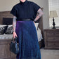 Person wearing a black turtleneck and blue pleated skirt in a bedroom setting.