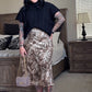 Woman standing in a bedroom wearing a black top and leopard print skirt.