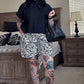Woman with tattoos standing in a bedroom wearing a black sweater and zebra print shorts.