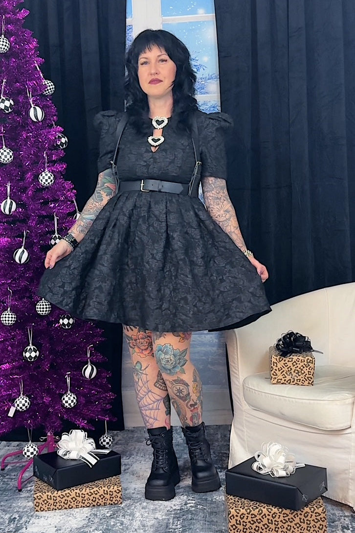Person wearing a black dress with tattoos standing next to a purple Christmas tree and gift boxes.