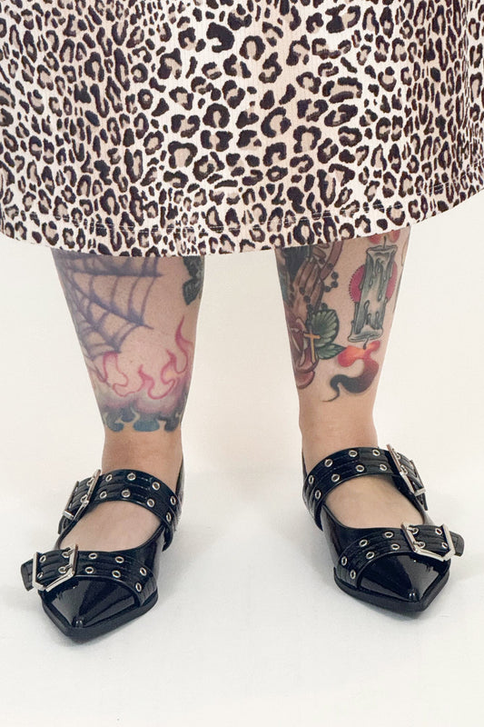 Person wearing black studded shoes with a leopard print skirt and tattoos on a white background