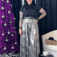 Woman in black top and silver sequin skirt standing next to a purple Christmas tree and gift boxes.