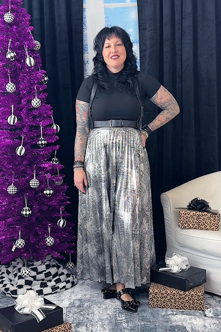 Woman in black top and silver sequin skirt standing next to a purple Christmas tree and gift boxes.