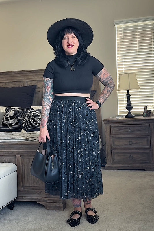 Woman in a black hat and black top with a star-patterned skirt standing in a bedroom.