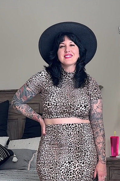 A woman wearing a leopard print pencil skirt with a seamless waistband, paired with a long-sleeve top, black hat, and carrying a black bag.
