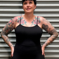 A tattooed woman with short dark hair stands in front of a gray metal roll-up door, wearing a black spaghetti-strap camisole top and leopard-print leggings.