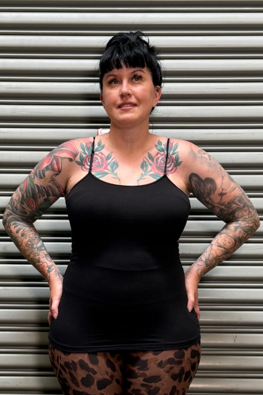A tattooed woman with short dark hair stands in front of a gray metal roll-up door, wearing a black spaghetti-strap camisole top and leopard-print leggings.