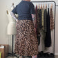 A person stands in a room wearing a leopard-print midi skirt with a dark t-shirt and black boots, facing a clothing rack with other garments. A dress form is visible to the left.