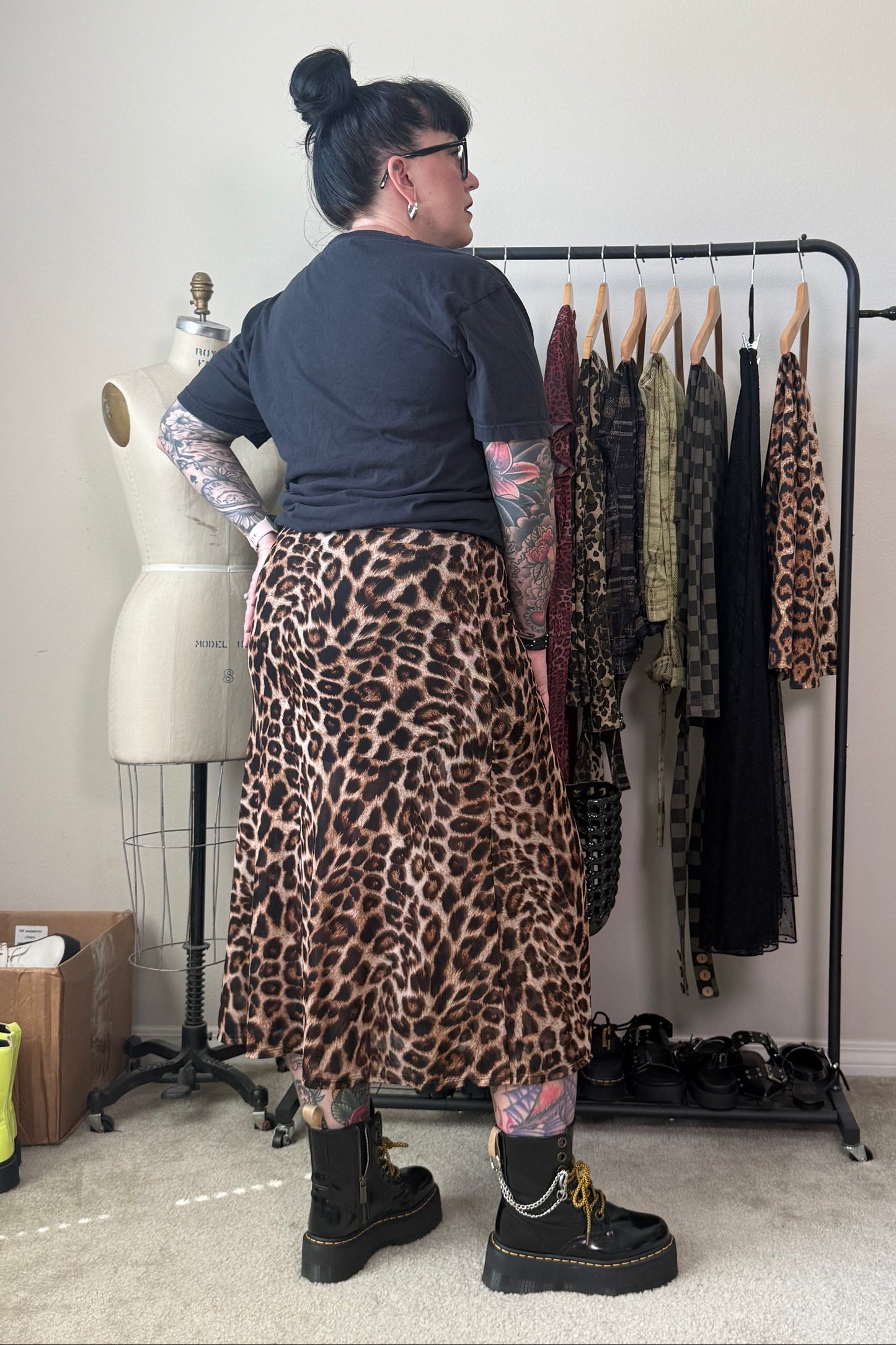 A person stands in a room wearing a leopard-print midi skirt with a dark t-shirt and black boots, facing a clothing rack with other garments. A dress form is visible to the left.