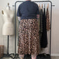 A person stands with their back to the camera wearing a leopard-print midi skirt and a dark t-shirt, standing in front of a clothing rack in a room with a dress form and shelves.